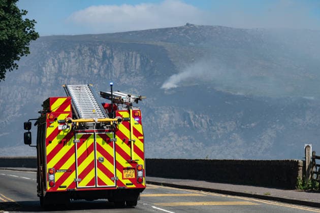 A Fire Engine drives past smouldering moorland close to Dove Stone Reservoir in Greater Manchester on 13 July 2026. Greater Manchester Police have arrested a woman in connection with a large moorland fire at Dove Stone Reservoir in Greenfield. The blaze, burning since Saturday, is under active investigation, with officers following leads regarding reports of fireworks nearby. The 20-year-old suspect remains in custody as the cause of the fire is yet to be officially determined. Oldham Greater Manchester England Dovestone Reservoir  Copyright: xMattxWilkinsonx
