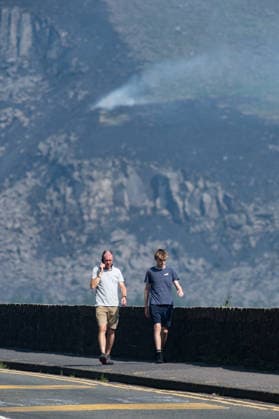Two men walk past smouldering moorland close to Dove Stone Reservoir in Greater Manchester on 13 July 2026. Greater Manchester Police have arrested a woman in connection with a large moorland fire at Dove Stone Reservoir in Greenfield. The blaze, burning since Saturday, is under active investigation, with officers following leads regarding reports of fireworks nearby. The 20-year-old suspect remains in custody as the cause of the fire is yet to be officially determined. Oldham Greater Manchester England Dovestone Reservoir  Copyright: xMattxWilkinsonx