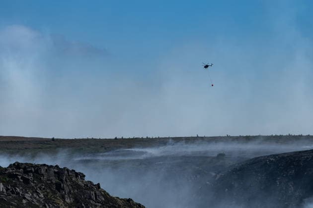 A helicopter carries water over smouldering moorland close to Dove Stone Reservoir in Greater Manchester on 13 July 2026. Greater Manchester Police have arrested a woman in connection with a large moorland fire at Dove Stone Reservoir in Greenfield. The blaze, burning since Saturday, is under active investigation, with officers following leads regarding reports of fireworks nearby. The 20-year-old suspect remains in custody as the cause of the fire is yet to be officially determined. Oldham Greater Manchester England Dovestone Reservoir  Copyright: xMattxWilkinsonx