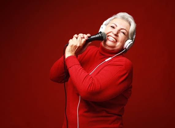 Porträt einer älteren Sängerin mit Kopfhörern und Mikrofon, die einen roten Pullover trägt, vor rotem Hintergrund Portrait of happy senior singer woman with headphones and mic wearing red sweater on red background (License=RF) ,model released, Symbolfoto Copyright: xZoonar.com RaisaxKanarevax 25327858 ,model released, Symbolfoto ,property released