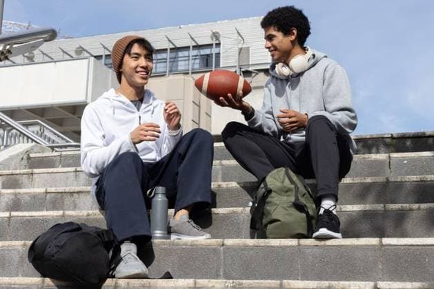 Diverse male friends sitting on concrete steps on campus passing football near backpacks headphones, model released, 08.08.2025 06:00:00, 7489166 Copyright: xIDS-260303_2xcreatorid:x0x 7489166