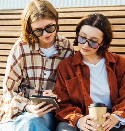 Zwei junge Studentinnen unterhalten sich an einem Sommertag, während sie auf einer Holzbank sitzen und eine Tasse Kaffee zum Mitnehmen in der Hand halten. Two young women chat while sitting on a wooden bench on a summer day with cups of coffee to go. (License=RF) ,model released, Symbolfoto Copyright: xZoonar.com RaisaxKanarevax 25334658 ,model released, Symbolfoto ,property released