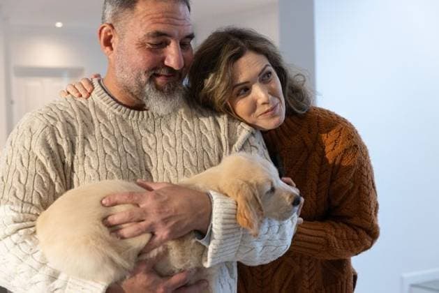 Couple holding light-colored puppy and leaning together in home hallway wearing cable-knit sweaters, model released, property released, 10.07.2025 06:00:00, 7009921 Copyright: xIDS-260525_1xcreatorid:x0x 7009921