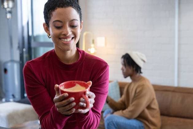 African American couple smiling, woman holding red mug in red sweater, man using smartphone at home, model released, property released, 29.08.2025 06:00:00, 8073776 Copyright: xIDS-260509_1xcreatorid:x0x 8073776