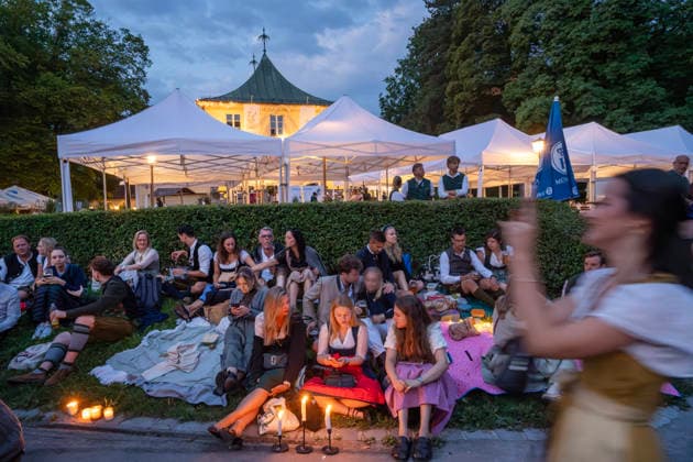 Kocherlball am Chinesischen Turm, Besucher haben es sich auf der Wiese niedergelassen, warten auf das Tanzvergnügen ab 6 Uhr, München, 19. Juli 2026 Deutschland, München, 19. Juli 2026, Biertrinken bei Kerzenschein, traditioneller Kocherlball am Chinesischen Turm, Englischer Garten, Besucher haben es sich auf der Wiese niedergelassen, warten auf das Tanzvergnügen ab 6 Uhr, manche Besucher sind schon um 3 Uhr früh im Biergarten, um noch einen Platz zu ergattern, die Menschen trinken ein Bier, essen eine Brotzeit, tragen Tracht, Tradition entstand im 19. Jahrhundert, als Hauspersonal, Dienstboten und KöchInnen in den frühen Morgenstunden vor Dienstbeginn gefeiert und getanzt haben, Tradition wurde ab 1989 wiederbelebt, traditionell am