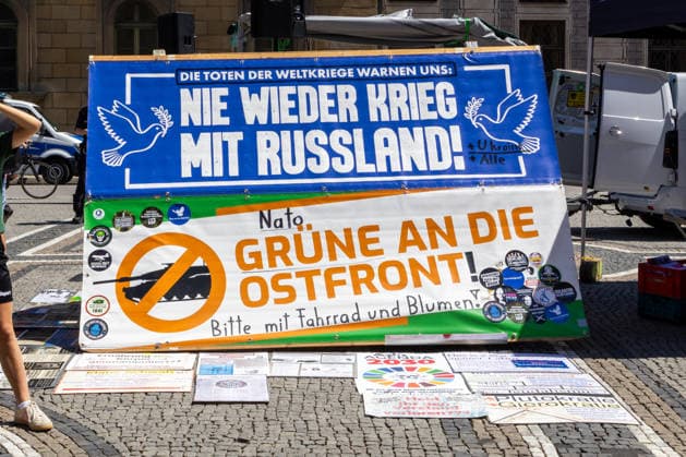 Munich, Bavaria, Germany July 25, 2026: Protesters demonstrate in downtown Munich against Chancellor Friedrich Merz (CDU) and the federal government™s policies. The rally is directed, among other things, against the government™s policies and social policies. Banners and signs read: The dead of the world wars warn us: Never again war with Russia; NATO and the Greens to the Eastern Front! Please bring bicycles and flowers *** Teilnehmer demonstrieren in der Münchner Innenstadt gegen Bundeskanzler Friedrich Merz (CDU) und die Politik der Bundesregierung. Die Kundgebung richtet sich unter anderem gegen die Regierungs- und Sozialpolitik. Banner bzw. Aufsteller mit Aufchrift: Die Toten der Weltkriege warnen uns: Nie wieder Krieg mi