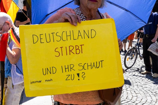 Munich, Bavaria, Germany July 25, 2026: Protesters demonstrate in downtown Munich against Chancellor Friedrich Merz (CDU) and the federal government™s policies. The rally is directed, among other things, against the government™s economic and social policies. An elderly woman holds a sign that reads: GERMANY IS DYING, AND YOU™RE STILL JUST STANDING BY AND WATCHING!? *** Teilnehmer demonstrieren in der Münchner Innenstadt gegen Bundeskanzler Friedrich Merz (CDU) und die Politik der Bundesregierung. Die Kundgebung richtet sich unter anderem gegen die Regierungs- und Sozialpolitik. Eine ältere Frau hält ein Plakat mit Aufschrift: DEUTSCHLAND STIRBT UND IHR SCHAUT IMMER NOCH ZU!?