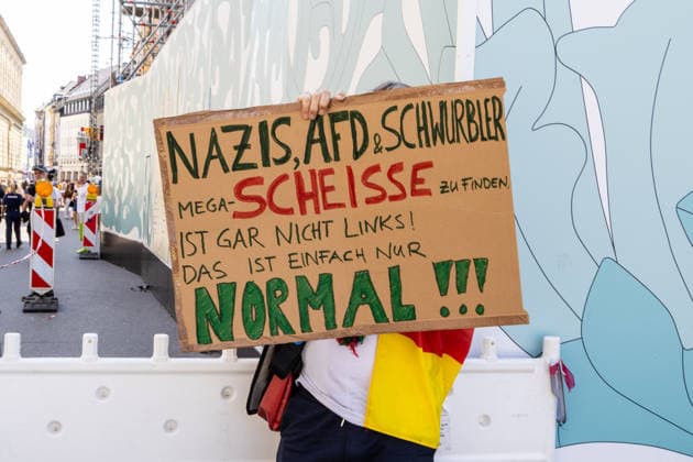 Munich, Bavaria, Germany July 25, 2026: Protesters demonstrate in downtown Munich against Chancellor Friedrich Merz (CDU) and the federal government™s policies. The rally is directed, among other things, against the government™s policies and social policies. A counter-protester standing on the sidelines of the demonstration holds a sign reading: NAZIS, AFD (Alternative for Germany), and SCHWURBLER THINKING THIS IS MEGA-CRAP ISN™T ˜LEFT-WING™ AT ALL! IT™S JUST NORMAL!!! *** Teilnehmer demonstrieren in der Münchner Innenstadt gegen Bundeskanzler Friedrich Merz (CDU) und die Politik der Bundesregierung. Die Kundgebung richtet sich unter anderem gegen die Regierungs- und Sozialpolitik. Ein Gegendemonstrant hält am Rand