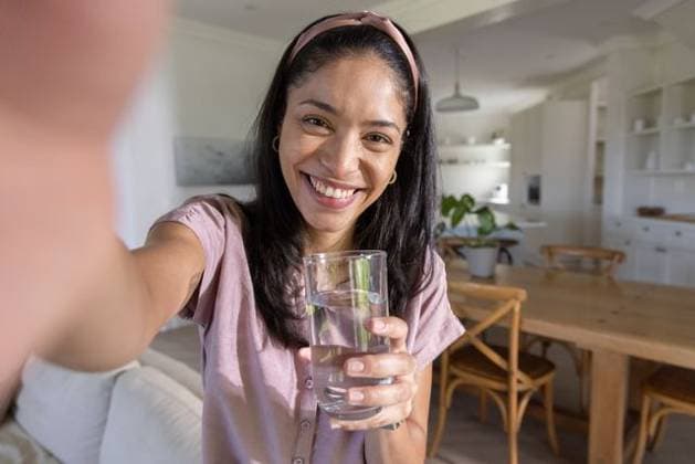 African woman wearing pink headband and shirt holding glass of water and taking selfie in kitchen, model released, property released, 20.02.2026 06:00:00, 16115865 Copyright: xIDS-260728xcreatorid:x0x 16115865