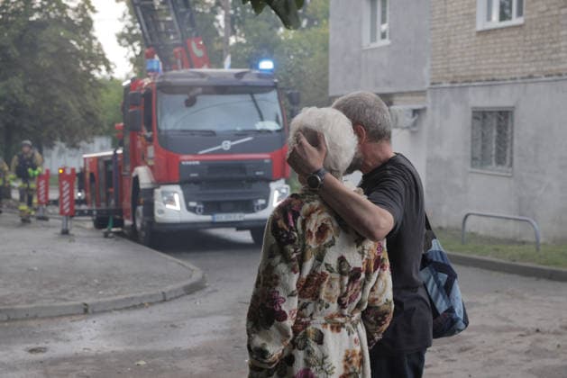 Shahed Drone Strike - Kharkiv A man consoles an elderly white-haired woman wrapped in a floral blanket as they stand before a Volvo aerial ladder fire truck with blue emergency lights activated following a russian Shahed drone strike that ignited the roof of a five-story residential building in the Novobavarski district of Kharkiv, Ukraine, on July 28, 2026, with all residents evacuated from the building, two people reported injured and received medical assistance as emergency crews worked to extinguish the blaze, and a firefighter in protective gear visible at left beside red safety barriers and hose lines stretched across the road. Photo by Independent Independent ABACAPRESS.COM Kharkiv Kharkiv Oblast Ukraine  Copyright: xTitovxYevhen ABACAx