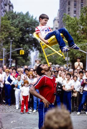 street performer holds chair and boy with his mouth in Washington Square Park in Manhattan, New York City, NY, USA in June 1982 *** A street performer holds a chair and a boy with his mouth in Washington Square Park in Manhattan, New York City, NY, USA, in June 1982