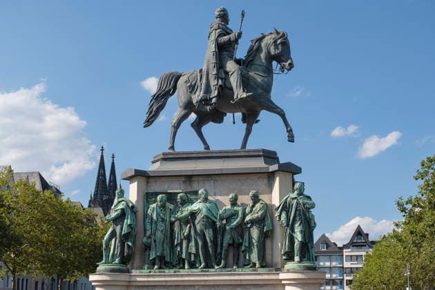 Das monumentale Reiterstandbild von König Friedrich Wilhelm III. von Preußen auf dem Heumarkt in der Kölner Altstadt. Das historische Bronzedenkmal zeigt den preußischen König hoch zu Ross, umgeben von bedeutenden Figuren der preußischen Geschichte am Sockel. Im Hintergrund sind bei blauem Sommerhimmel die Spitzen des Kölner Doms zu sehen. Das Denkmal steht symbolisch für die preußische Geschichte des Rheinlands, Denkmalschutz, Kunstgeschichte, Stadtarchitektur und den Tourismus in Köln. *** The monumental equestrian statue of King Frederick William III of Prussia located at Heumarkt in the old town of Cologne, Germany. The historic bronze monument depicts the Prussian king on horseback, surrounded by prominent figures of Prussian history a GMS20498