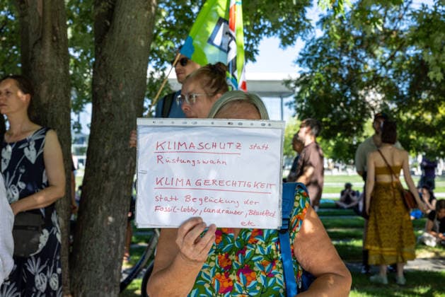Fridays for Future Berlin - Hitzedemo. Deutschland, Berlin am 6. August 2026: Eine Frau hält ein kleines Schild hoch Klimaschutz statt Rüstungswahn. Klimagerechtigkeit statt Beglückung der Fossil-Lobby, Landräuber, Blackrock etc. Vor dem Kanzleramt protestieren Klimaaktivistinnen und Klimaaktivisten unter dem Motto Klimakrise eskaliert, Kanzler schweigt?!. Sie verlangen verstärkte Klimaschutzmaßnahmen, einen schnellen politischen Hitzeschutz sowie einen konsequenten Ausstieg aus fossilen Energien. *** Fridays for Future Berlin Heat Protest, Germany, Berlin, August 6, 2026. A woman holds up a small sign reading: Climate Protection Instead of the Arms Race; Climate Justice Instead of Catering to the Fossil Fuel Lobby, Land Gr