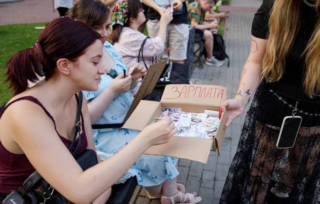 An activist holding a box signed Salary offers souvenir banknotes to those present as part of an ironic performance during an open-ended peaceful protest against the dismissal of Mykhailo Fedorov from the post of Ukraine’s Defence Minister in Ivana Franka Square outside the Ivan Franko National Academic Drama Theatre, close to the Office of the President, Kyiv, Ukraine, August 6, 2026. Large-scale cardboard rallies in support of former Defence Minister Mykhailo Fedorov have been held in Kyiv and other cities across Ukraine for 22 consecutive days. On 14 July 2026, the Ukrainian Parliament accepted the resignation of Prime Minister Yuliia Svyrydenko, leading to the resignation of the Cabinet. On 15 July 2026, President Volodymyr Zelensky  Copyright: xMariannaxKotykx
