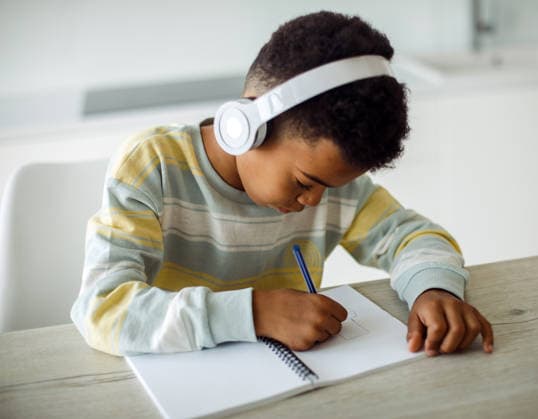 Ein Junge gemischter Herkunft schreibt in ein Notizbuch und hört über Kopfhörer Musik A mixed race boy writes in a notebook and listens to music on headphones at home. (License=RF) ,model released, Symbolfoto Copyright: xZoonar.com RaisaxKanarevax 25358179 ,model released, Symbolfoto ,property released