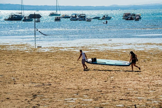 Zwei Wassersportler tragen ihr großes Stand Up Paddle Board durch die breite trockengefallene Uferzone zum Wasser, Wasserburg, August 2026 Deutschland, Wasserburg, August 2026, Niedrigwasser am Bodensee, zwei Wassersportler tragen ihr großes Stand Up Paddle Board durch die breite trockengefallene Uferzone zum Wasser, durch Trockenheit freigelegte Uferzone am Bodensee,, die aktuelle Trockenheit hat den Pegelstand stark sinken lassen, die Boote liegen weit draußen, die Schifffahrt ist eingeschränkt, Niedrigwasser, Wasserstand sehr niedrig, zu wenig Regen, schönes Wetter, Hochsommer, Hitzewelle mit Temperaturen bis 33 Grad, Sommer, Landkreis Lindau, Bayern, *** Two water sports enthusiasts carry their large stand-up paddleboard across th