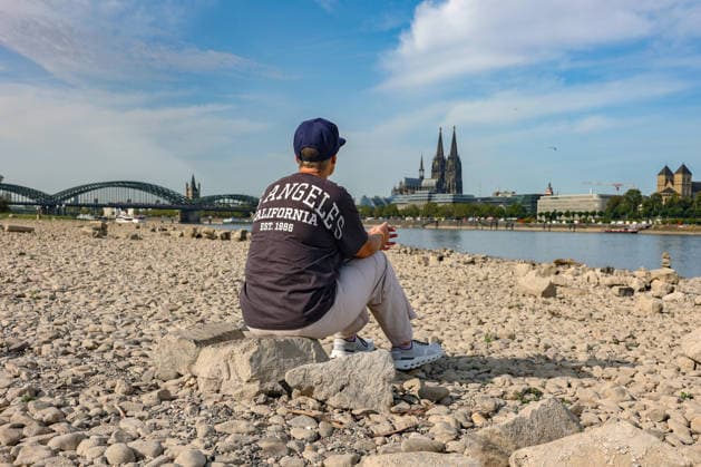 Sommer im Rheinland. Blick auf den Rhein bei Koeln (Köln) - Deutz. Der Rhein in Koeln (Köln) fuehrt (führt) Niedrigwasser aufgrund anhaltender Trockenheit. Im Hintergrund die Hohenzollernbruecke (Hohenzollernbrücke) (links) und der Koelner (Kölner) Dom (rechts). Im Vordergrund sitzt eine Frau auf einem Stein und schaut auf den Rhein. Sommer im Rheinland am 09.08.2026 in Koeln (Köln) Deutschland. *** Summer in the Rhineland: View of the Rhine near Cologne (Deutz). The Rhine in Cologne is at low water levels due to persistent drought. In the background, the Hohenzollern Bridge is on the left and Cologne Cathedral is on the right. In the foreground, a woman is sitting on a rock, looking out at the Rhine. Summer in the Rhineland on Augus