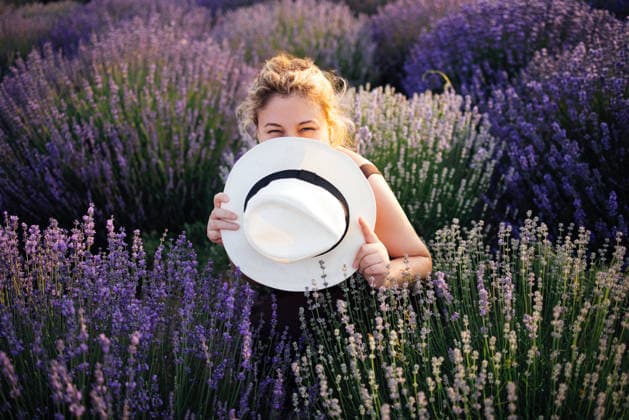 Eine junge Frau mit lockigem Haar hält spielerisch einen weißen Hut in einem Lavendelfeld in der Hand, umgeben von leuchtend violetten Blüten unter einem strahlend blauen Himmel bei sonnigem Wetter Young woman with curly hair smiles while holding a white hat in a lavender field, surrounded by blooming purple flowers and a bright blue sky (License=RF) ,model released, Symbolfoto Copyright: xZoonar.com DashaxPetrenkox 25405397 ,model released, Symbolfoto ,property released