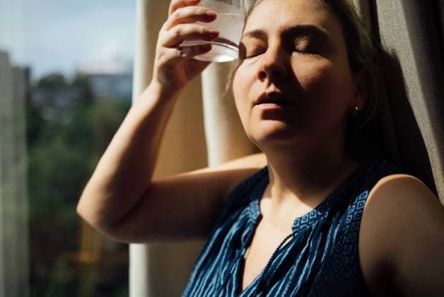 Eine Frau mit hellbraunen Haaren, die sich ein Glas Wasser an die Stirn hält, die Augen geschlossen, steht an einem Fenster, wobei natürliches Licht ihr Gesicht und ihre Umgebung beleuchtet Woman with light brown hair holds a glass of water against her forehead, eyes closed, near a window with soft natural light illuminating the scene (License=RF) ,model released, Symbolfoto Copyright: xZoonar.com DashaxPetrenkox 25405428 ,model released, Symbolfoto ,property released