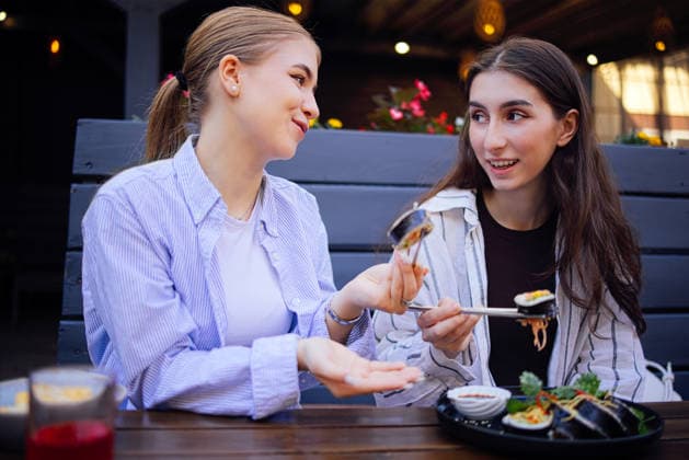 Zwei junge Frauen genießen Sushi in einem Restaurant im Freien; die eine hält Sushi mit Essstäbchen in der Hand, während die andere gestikuliert; im Hintergrund sind farbenfrohe Blumen und eine warme Beleuchtung zu sehen Two young women share sushi at an outdoor dining area, one using chopsticks to pick up sushi while the other gestures, with flowers and warm lighting around (License=RF) ,model released, Symbolfoto Copyright: xZoonar.com DashaxPetrenkox 25405446 ,model released, Symbolfoto ,property released