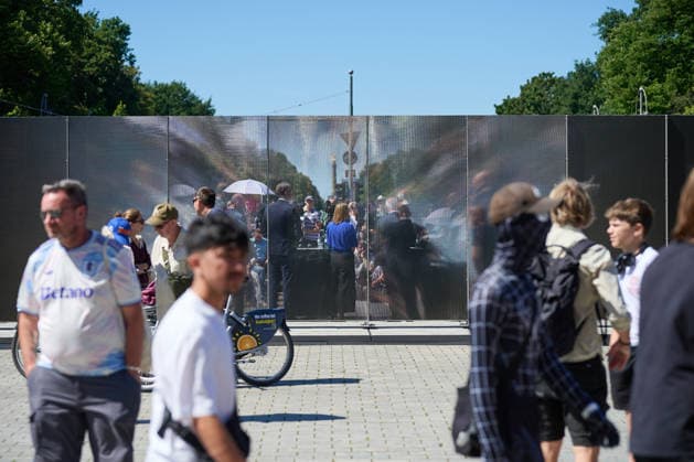 Ein temporaerer Gedenkort am Brandenburger Tor soll zum 65. Jahrestag des Mauerbaus an die deutsche Teilung erinnern (Foto vom 12.08.2026, im Bild: Menschen vor und hinter der Installation). Die rund drei Meter hohe und 30 Meter lange Installation des Kuenstlers Hubertus Hamm auf dem Platz des 18. Maerz soll an die einstigen Grenzanlagen erinnern. Je nach Perspektive wirkt die Wand aus Aluminium lichtundurchlaessig oder transparent. (Siehe epd-Meldung vom 12.08.2026) EDITORIAL USE ONLY *** A temporary memorial site at the Brandenburg Gate is intended to commemorate the division of Germany on the 65th anniversary of the construction of the Berlin Wall. Photo taken on August 12, 2026, showing people in front of and behind the installation. Th Copyright: epd-bild ChristianxDitsch D26D0812MauerbauInstallation123658