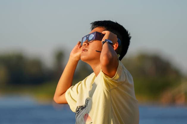 Altenrhein, Bodensee, Schweiz: Junge Person beobachtet mit einer Sonnenfinsternisbrille den Himmel und hält die Schutzbrille mit beiden Händen fest. Seitliches Porträt im warmen Abendlicht. Sonnenfinsternis, partielle Sonnenfinsternis, Sofi, Astronomie, Sonne, Himmelsbeobachtung, Sonnenfinsternisbrille, Augenschutz, Naturphänomen, Wissenschaft, Porträt, Symbolbild, Altenrhein, Bodensee, St. Gallen, Schweiz. *** Altenrhein, Lake Constance, Switzerland A young person observes the sky through solar eclipse glasses, holding the protective glasses with both hands. Side profile in the warm evening light. Solar eclipse, partial solar eclipse, Sofi, astronomy, Sun, sky observation, solar eclipse glasses, eye protection, natural phenomenon, Copyright: xJOERANxSTEINSIEKx