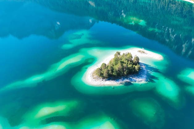 Wetterfeature, Morgenstille am Eibsee: Unterhalb der Zugspitze liegt der See bei Grainau nahe Garmisch-Partenkirchen in den frühen Stunden noch ruhig und nahezu unberührt. Doch an heißen Sommertagen ändert sich das Bild häufig schon ab Mittag. Dann strömen zahlreiche Ausflügler und Urlauber an den durch soziale Medien weltweit bekannt gewordenen Bergsee. Der zunehmende Massentourismus sorgt regelmäßig für einen regelrechten Besucheransturm. Unter dem hohen Verkehrsaufkommen, zugeparkten Straßen und dem großen Andrang leiden zunehmend auch die Anwohner in Grainau und rund um den Eibsee. (Aufnahme mit einer Drohne) Grainau Bayern Deutschland Grainau *** Weather Feature, Morning Stillness at Eibsee. Nestled below the Zugspitze near Copyright: x xonw-imagesx xMariusxBullingx