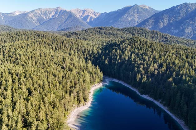Wetterfeature, Morgenstille am Eibsee: Unterhalb der Zugspitze liegt der See bei Grainau nahe Garmisch-Partenkirchen in den frühen Stunden noch ruhig und nahezu unberührt. Doch an heißen Sommertagen ändert sich das Bild häufig schon ab Mittag. Dann strömen zahlreiche Ausflügler und Urlauber an den durch soziale Medien weltweit bekannt gewordenen Bergsee. Der zunehmende Massentourismus sorgt regelmäßig für einen regelrechten Besucheransturm. Unter dem hohen Verkehrsaufkommen, zugeparkten Straßen und dem großen Andrang leiden zunehmend auch die Anwohner in Grainau und rund um den Eibsee. (Aufnahme mit einer Drohne) Grainau Bayern Deutschland Grainau *** Weather Feature, Morning Stillness at Eibsee. Nestled below the Zugspitze near Copyright: x xonw-imagesx xMariusxBullingx