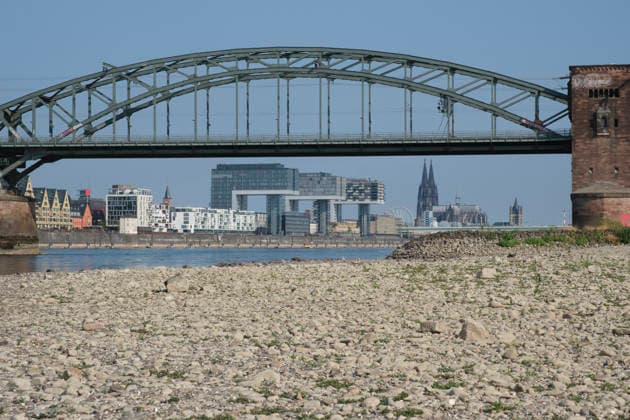Eine bodennahe Aufnahme zeigt das weitläufige, freigelegte Kiesel- und Steinufer des Rheins bei extremem sommerlichem Niedrigwasser. Im unscharfen Hintergrund erstreckt sich die stählerne Fachwerk-Konstruktion der historischen Südbrücke mit ihren markanten Wehrtürmen aus Backstein. Dahinter zeichnet sich die Kölner Skyline mit den modernen Kranhäusern im Rheinauhafen und den entfernten Spitzen des Kölner Doms unter einem klaren, blauen Himmel ab. *** A low-angle shot captures the wide, exposed pebble and stone bank of the Rhine during extreme summer low water levels. In the blurred background, the steel truss structure of the historic Südbrücke stands out with its distinctive brick defense towers. Behind it, the Cologne skyline features the 20260816-DSCF2123