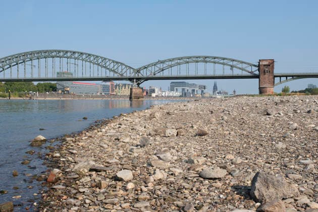 Eine weitwinklige Totalansicht zeigt den stark zurückgewichenen Flusslauf des Rheins mit einem weitläufig ausgetrockneten Kiesel- und Steinufer bei extremem sommerlichem Niedrigwasser. Die historische, stählerne Südbrücke überspannt den Fluss mit ihren charakteristischen Fachwerkbögen und dem markanten Wehrturm aus rotem Backstein. Im Hintergrund spiegelt sich das gegenüberliegende Rheinufer mit der Kölner Skyline, den Kranhäusern im Rheinauhafen und den entfernten Türmen des Kölner Doms unter einem wolkenlosen, blauen Himmel. *** A wide-angle view captures the significantly receded riverbed of the Rhine, exposing a vast dried-out pebble and stone bank during extreme summer low water levels. The historic steel Südbrücke spans the river with 20260816-DSCF2131