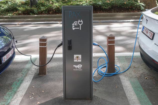 Zwei Elektrofahrzeuge sind über Ladekabel mit einer anthrazitfarbenen Ladesäule von TotalEnergies am Straßenrand verbunden. Auf der linken Seite ist das Heck eines blauen Fahrzeugs zu sehen, während rechts ein weißes Tesla-Modell mit belgischem Kennzeichen lädt. Die Ladesäule im Zentrum weist neben dem offiziellen Logo und einem E-Auto-Symbol ein handgemaltes Graffiti-Wort PAX sowie einen Aufkleber mit der Aufschrift Green Washing! auf.