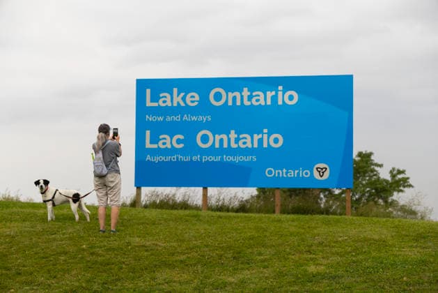 (260830) -- HAMILTON, Aug. 30, 2026 -- A woman takes photos of an English-French bilingual sign reading Lake Ontario Now and Always at a lakeside conservation area in Hamilton, Ontario, Canada, on Aug. 30, 2026. Canadian Prime Minister Mark Carney said Thursday that the name Lake Ontario carries a long history and that Canadians will always use this name regardless of external changes. The Canadian prime minister made the remarks after U.S. President Donald Trump signed an executive order earlier on Thursday, directing the Department of the Interior to rename the lake Lake America, as trade tensions between the United States and Canada have been escalating. (Photo by Xinhua) CANADA-ONTARIO-HAMILTON-LAKE ONTARIO-NAME ZouxZheng 