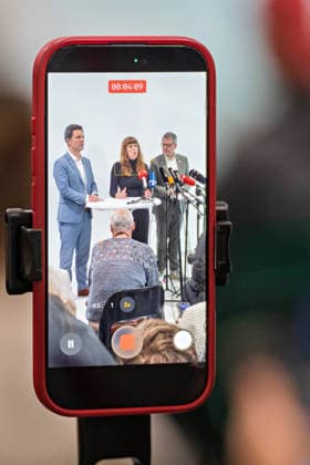 Berlin, 08.10.2026: Auf dem Display eines Smartphones sind Steffen Krach (SPD, l.), Kerstin Wolter-Ehling (Die Linke, M.) und Werner Graf (Bündnis 90 Die Grünen, r.) beim gemeinsamen Pressestatement nach Abschluss der Vorgespräche ihrer Parteien im Foyer des Hauses der Statistik zu sehen. Pressestatement von Linke, Grünen und SPD in Berlin