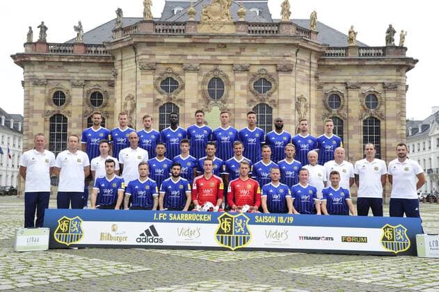 Das Team des 1. FC Saarbrücken am Donnerstag (12.07.2018) vor der Ludwigskirche in Saarbrücken. Im Bild: Obere Reihe (v.l.) Marco Holz ( 14), Macel Carl ( 22), Steven Zellner ( 5), Alexandre Mendy ( 4), Benjamin Kessel ( 15), Oliver Oschkenat ( 3), Nino Miotke ( 28), José Pierre Vunguidica ( 9), Sebastian Jacob ( 24) und Marco Kehl-Gomez. Mittlere Reihe: (v.l.) Robert Roelofsen (Co-Trainer), Dirk Lottner (Cheftrainer), Christoph Fuhr (Athletiktrainer), Heinz Böhmann (Torwart-Trainer), Gillian Jurcher ( 21), Lukas Quirin ( 31), Markus Obernosterer ( 10), Manuel Zeitz ( 8), Tobias Jänicke ( 25), Pierre Fassnacht ( 29), Helmut Schwan (Betreuer), Rüdiger Schmid (Zeugwart), Paulo da Palma (Physiotherapeut) und Frank Kackert (Torwart-Trainer). Untere Reihe: (v.l.) Fatih Köksal ( 27), Sascha Wenninger ( 19), Fanol Perdedaj ( 17), Ricco Cymer ( 20), Daniel Batz ( 1), Martin Dausch ( 7), Markus Mendler ( 11) und Mario Müller ( 23).