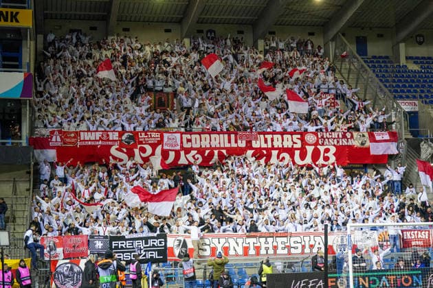 Belgien: Fussball Europa League 2025 - 2026, Achtelfinale - KRC Genk vs SC Freiburg - Cegeka Arena (Genk): Genk, Belgien, Europa: Fanblock des SC Freiburg im Cegeka Arena Stadion während eines UEFA Europa League Spiels gegen KRC Genk, Auswärtsfans mit rot-weißen Fahnen, Ultras, Banner Corrillo Ultras, Synthesia, Fight Fascism, Freiburg Fankultur, Gästetribüne, Europapokal, internationaler Vereinsfussball, Stadionatmosphäre, Flutlichtspiel, Fussballfans, Supporters, Tribüne, Matchday, UEFA Europa League. Copyright: xJOERANxSTEINSIEKx