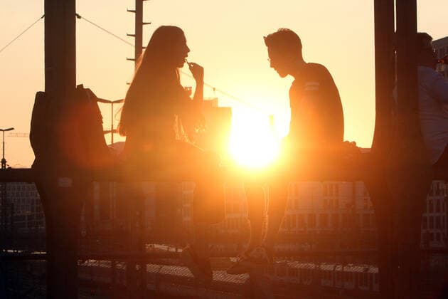 Ein Paar sitzt im Gegenlicht der untergehenden Sonne auf einem Träger der Hackerbrücke in München und unterhält sich, Bayern, Deutschland.