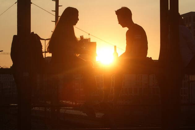 Ein Paar sitzt im Gegenlicht der untergehenden Sonne auf einem Träger der Hackerbrücke in München und unterhält sich, Bayern, Deutschland.