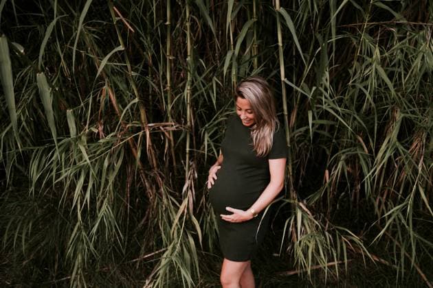 Happy pregnant woman in casual dress stroking belly while standing on green trees wall in sunny summer day with blue sky, Model released Copyright: xSantixMiquelx MR