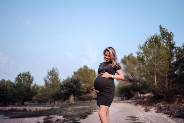 Happy pregnant woman in casual dress stroking belly while standing on pathway in park with green trees in sunny summer day with blue sky, Model released Copyright: xSantixMiquelx MR