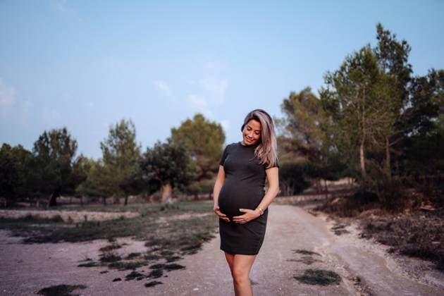 Happy pregnant woman in casual dress stroking belly while standing on pathway in park with green trees in sunny summer day with blue sky, Model released Copyright: xSantixMiquelx MR