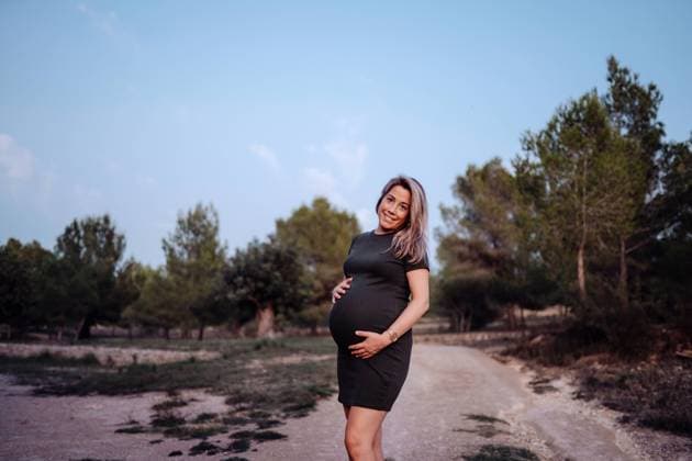 Happy pregnant woman in casual dress stroking belly while standing on pathway in park with green trees in sunny summer day with blue sky, Model released Copyright: xSantixMiquelx MR
