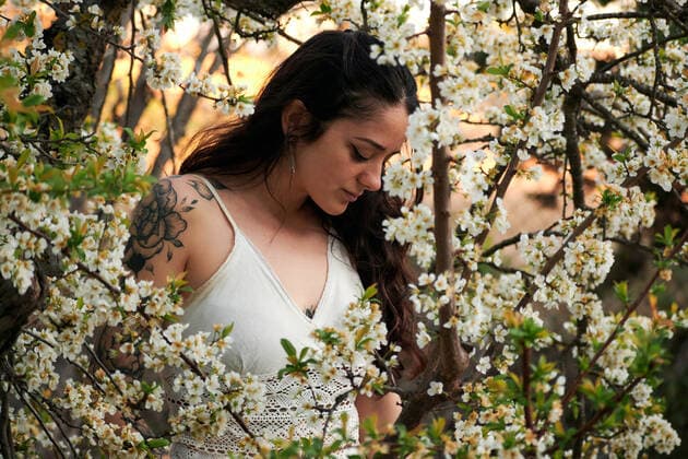 Young female with tattooed arm wearing white dress and standing in flowers of tree looking down, Model released, Property released Copyright: xCristianxBlazquezx MR
