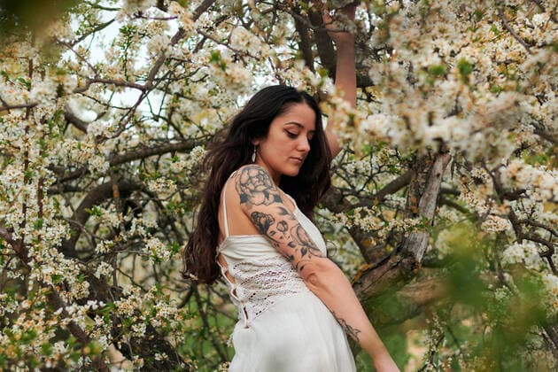 Young female with tattooed arm wearing white dress and standing in flowers of tree looking down, Model released, Property released Copyright: xCristianxBlazquezx MR