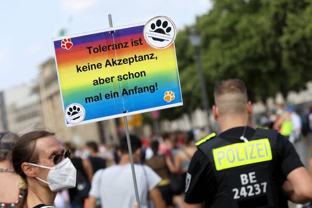 Berliner CHRISTOPHER STREET DAY 2021 - CSD - Am 24. Juli 2021 zieht der CSD in Berlin unter dem Motto SAVE OUR COMMUNITY Ð SAVE YOUR PRIDE zum 43. Mal durch die Hauptstadt. Demoteilnehmer mit weißer Mundschutzmaske und Sonnenbrille trägt Protestschild in Regenbogenfarben mit der Aufschrift: Toleranz ist keine Akzeptanz, aber schon mal ein Anfang! - PUPPLAY GERMANY - Rechts im Bild: Polizei zur Sicherung der Demonstration Die bunte CSD-Parade startete in diesem Jahr als Demonstration an der Leipziger Straße, führte über den Potsdamer Platz, vorbei am Denkmal für die im Nationalsozialismus verfolgten Homosexuellen zum Brandenburger Tor und über die Siegessäule zur Urania. Aufgrund der Corona-Pandemie fand die Demonstration ausschließlich unt