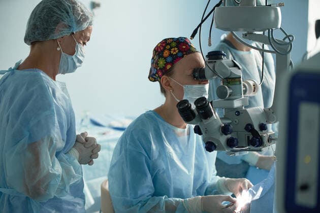 Adult female doctor in sterile mask and ornamental medical cap looking through surgical microscope against crop coworker in hospital, Model released SergeyMironov_Surgery_18.jpg Copyright: xSergeyxMironovx MR