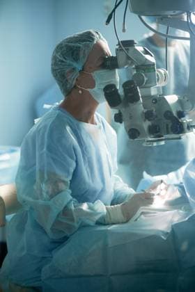 Adult female doctor in sterile mask and ornamental medical cap looking through surgical microscope against crop coworker in hospital, Model released SergeyMironov_Surgery_22.jpg Copyright: xSergeyxMironovx MR