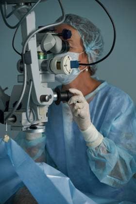 Adult female doctor in sterile mask and ornamental medical cap looking through surgical microscope against crop coworker in hospital, Model released SergeyMironov_Surgery_23.jpg Copyright: xSergeyxMironovx MR
