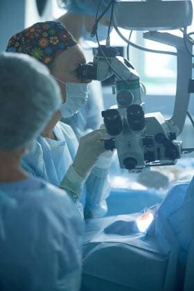 Adult female doctor in sterile mask and ornamental medical cap looking through surgical microscope against crop coworker in hospital, Model released SergeyMironov_Surgery_3.jpg Copyright: xSergeyxMironovx MR