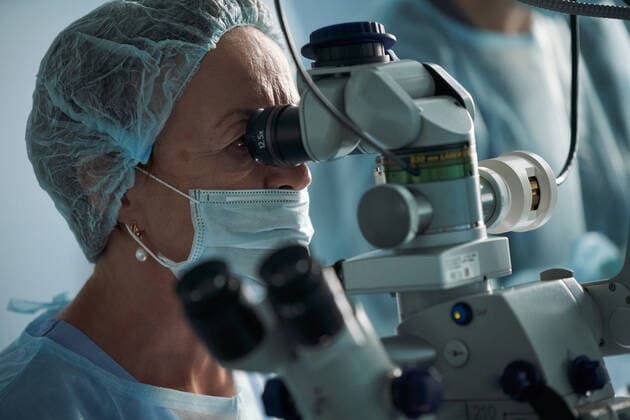 Adult female doctor in sterile mask and ornamental medical cap looking through surgical microscope against crop coworker in hospital, Model released SergeyMironov_Surgery_30.jpg Copyright: xSergeyxMironovx MR