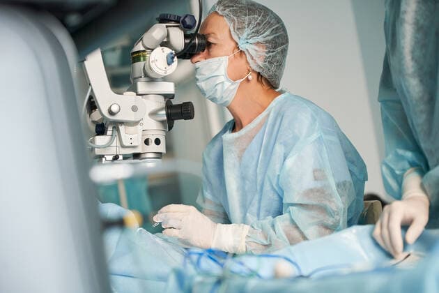 Adult female doctor in sterile mask and ornamental medical cap looking through surgical microscope against crop coworker in hospital, Model released SergeyMironov_Surgery_32.jpg Copyright: xSergeyxMironovx MR